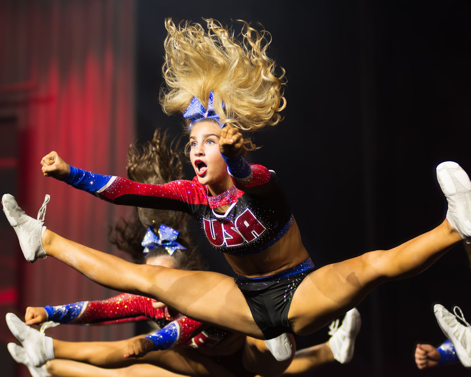 Cheer athlete mid toe-touch jump, hair flying, in front of a red stage