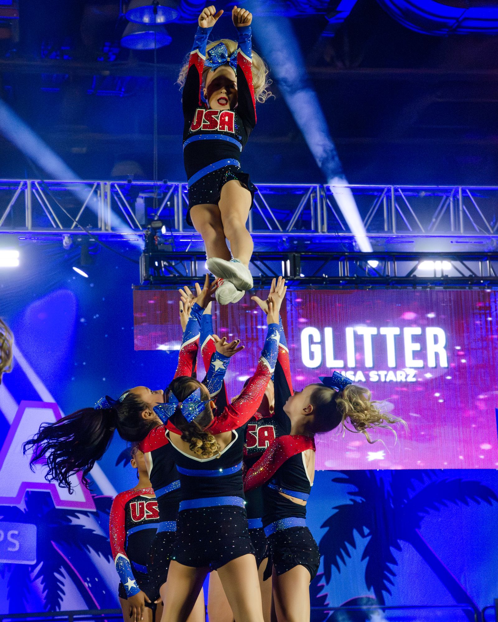 Tall cheer stunt with the flyer high above her group under blue stage lights