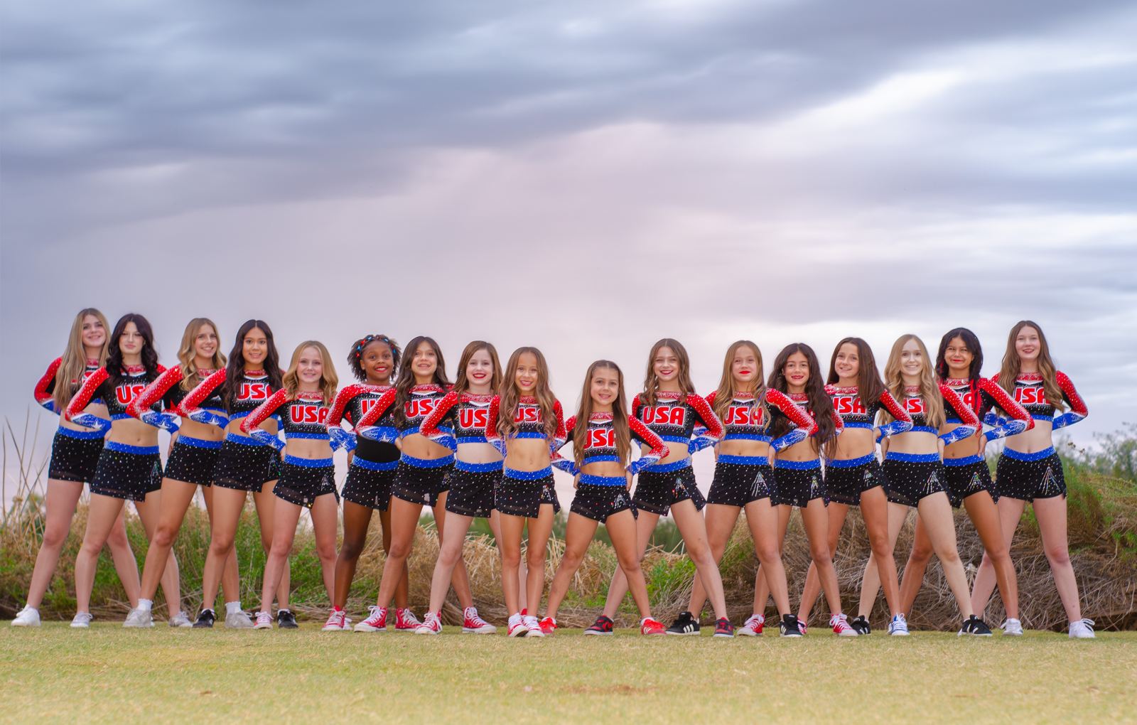 Cheer team in red, white, and blue uniforms lined up on a grassy field under a stormy sky