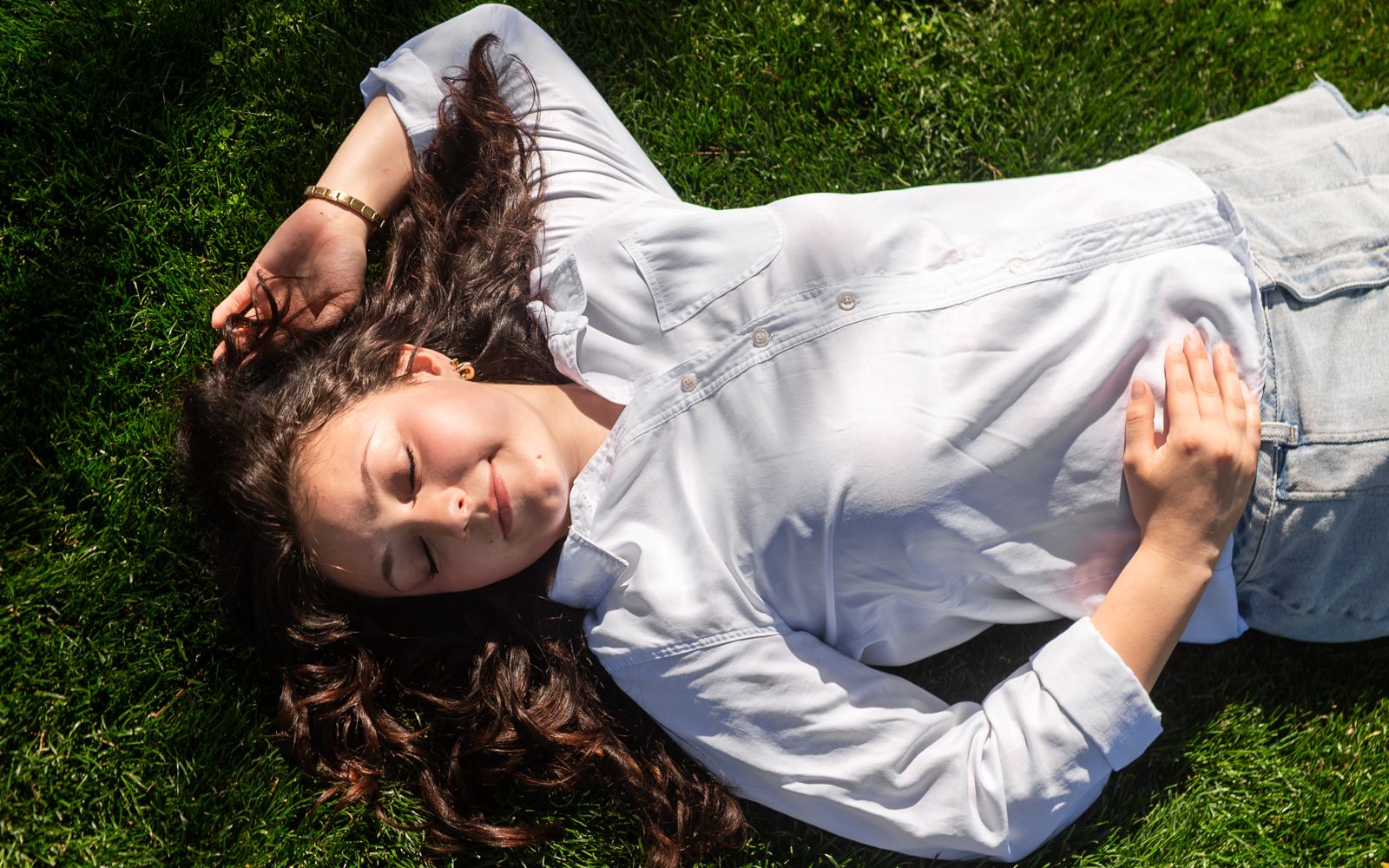 Teen in a white button-up shirt lying in the grass with her eyes closed, smiling in the sun