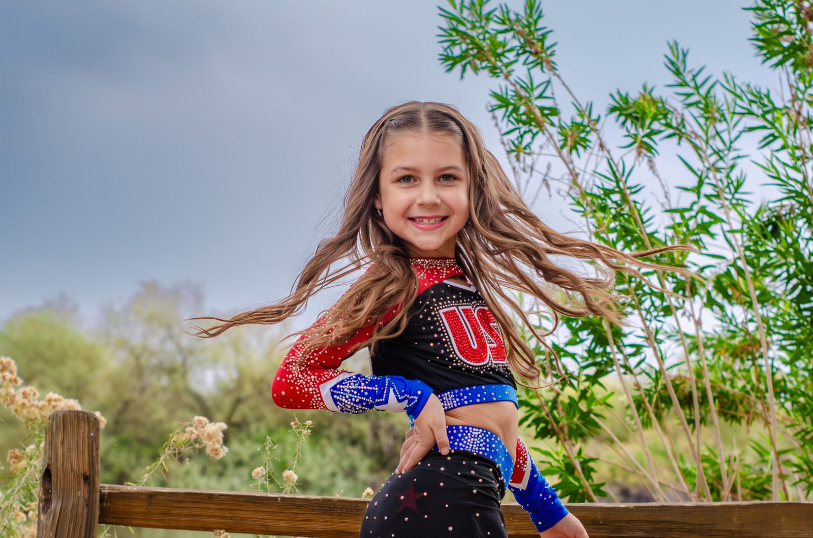 Smiling girl in a cheer uniform, hair flying, beside a wooden fence and greenery