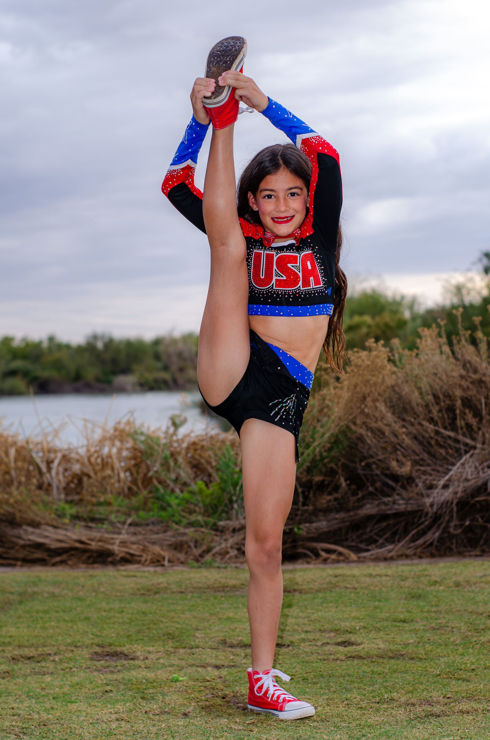 Girl in a cheer uniform holding a standing split on the grass by a lake